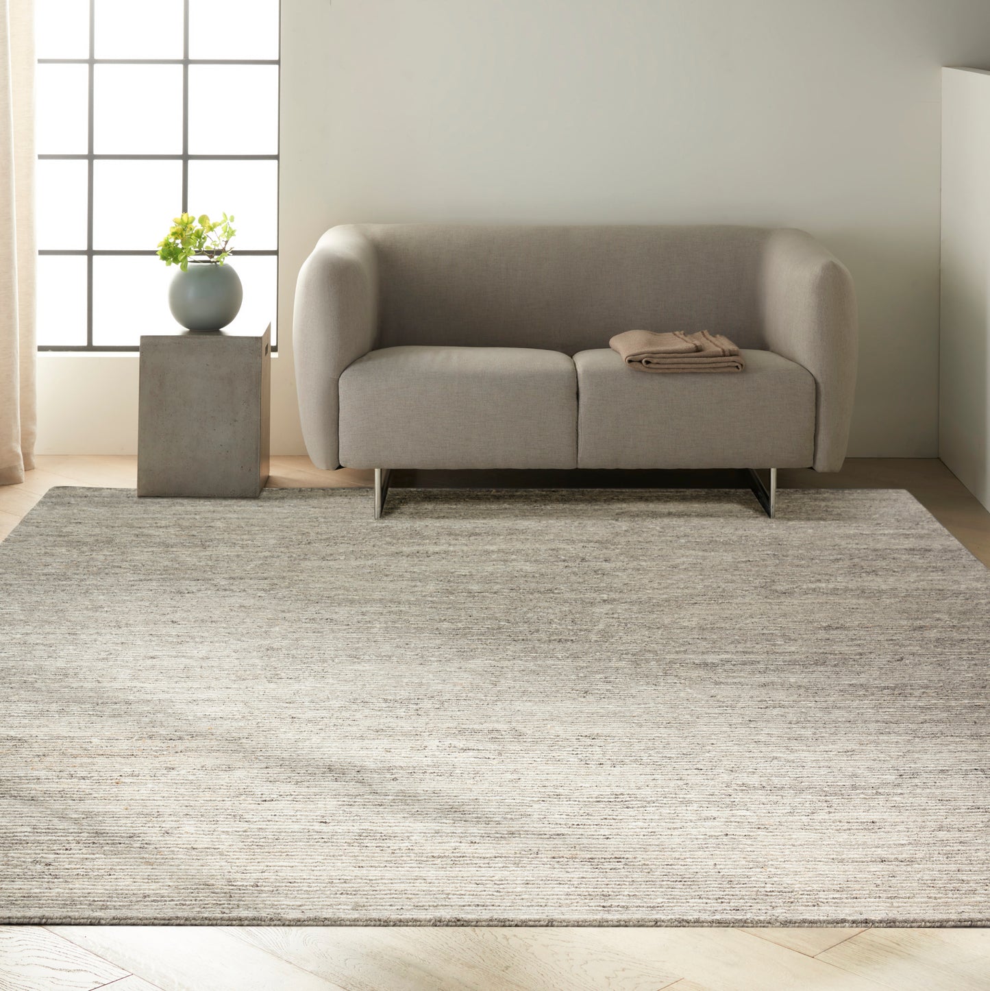 Modern light-gray sofa with rounded arms and metal legs, on a textured beige rug; concrete side table with plant.