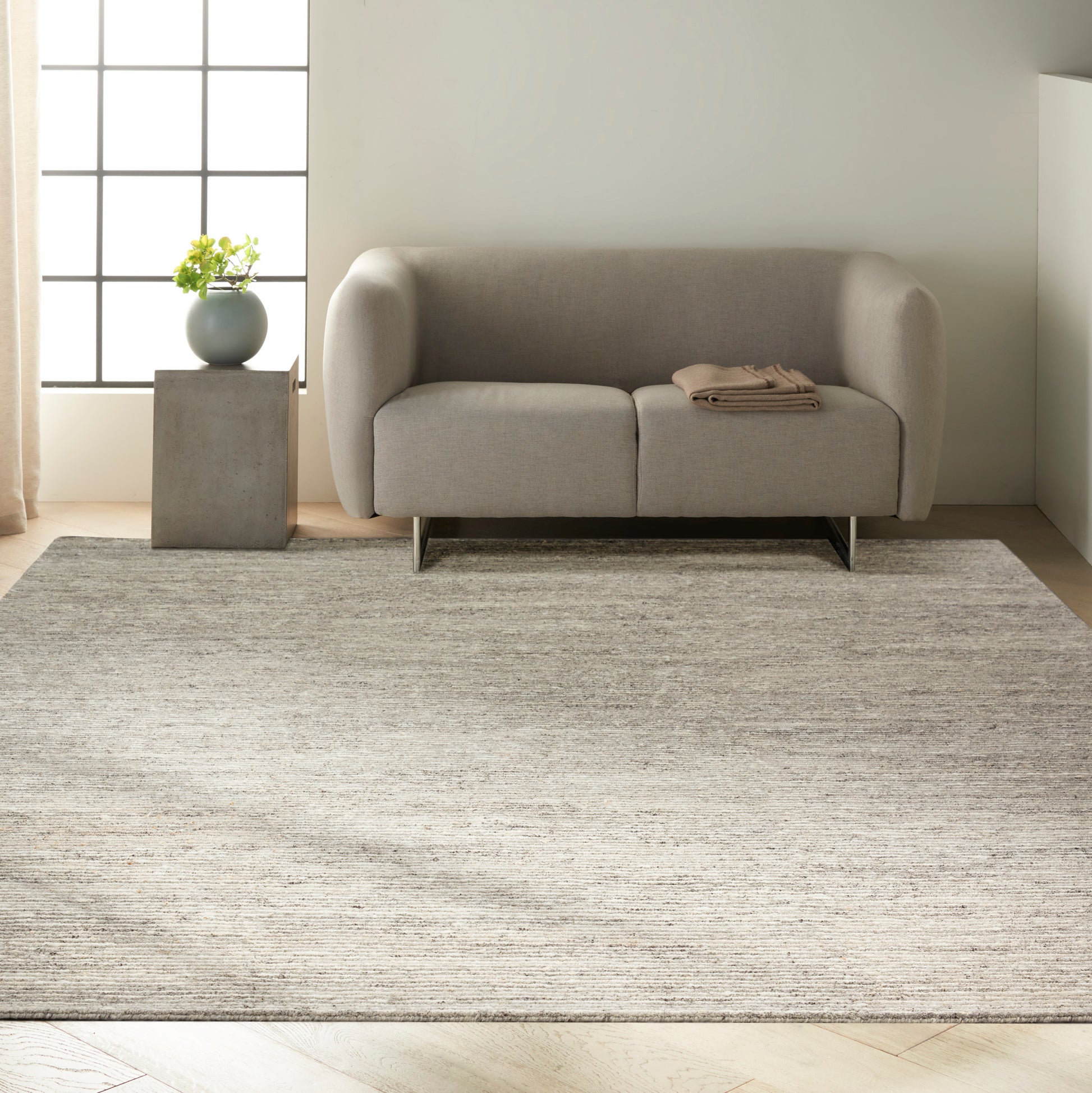 Modern light-gray sofa with rounded arms and metal legs, on a textured beige rug; concrete side table with plant.