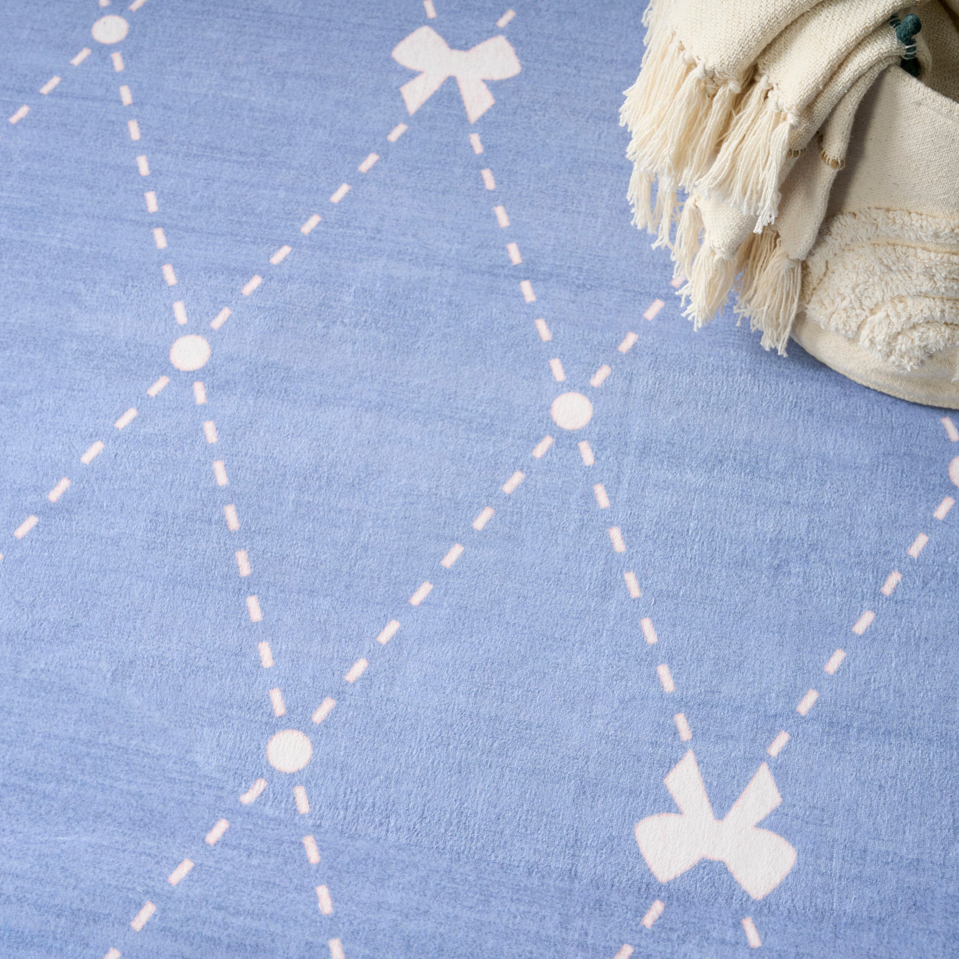Blue rug with white dashed diamond lattice and bow motifs; beige fringed throw in the top-right corner.