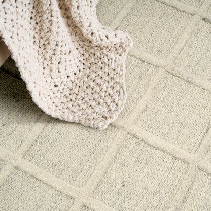 Cream chunky knit blanket draped over a light beige rug with square geometric pattern.