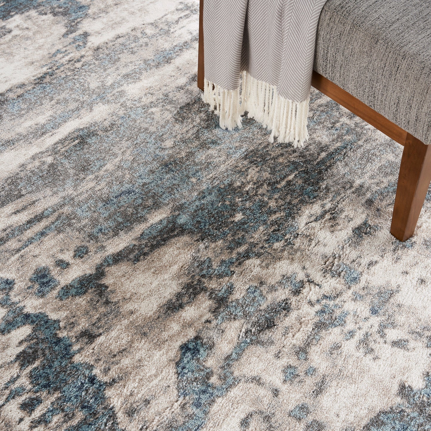 Abstract blue-gray rug with cream and navy tones under a grey sofa with wooden legs and fringe.
