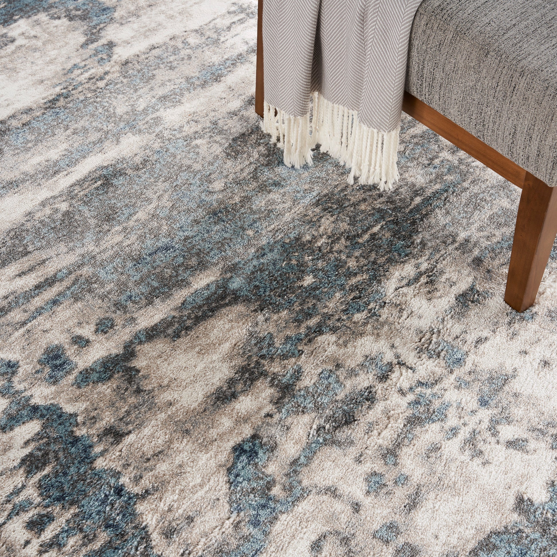 Abstract blue-gray rug with cream and navy tones under a grey sofa with wooden legs and fringe.