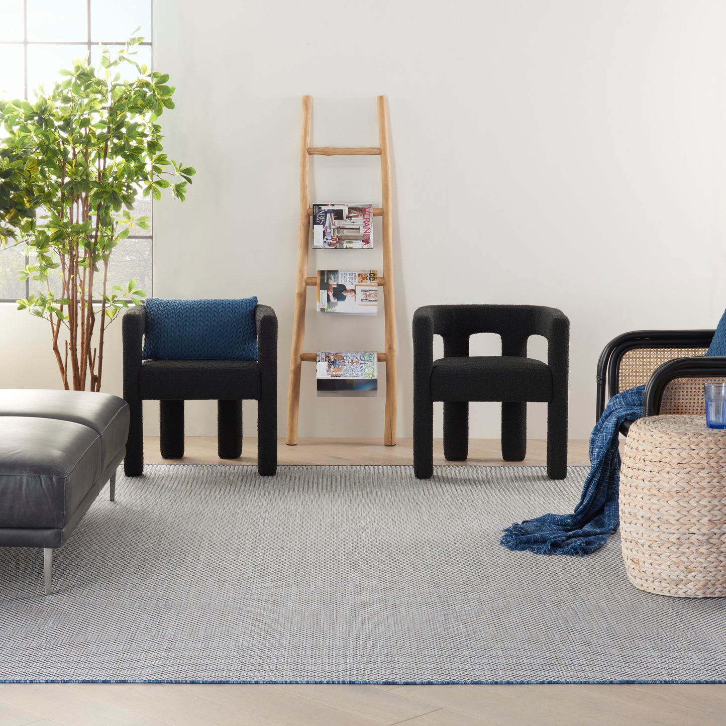 Bright minimalist living room with two black lounge chairs, a wooden ladder shelf, a wicker basket with blue throw, plant, gray rug.