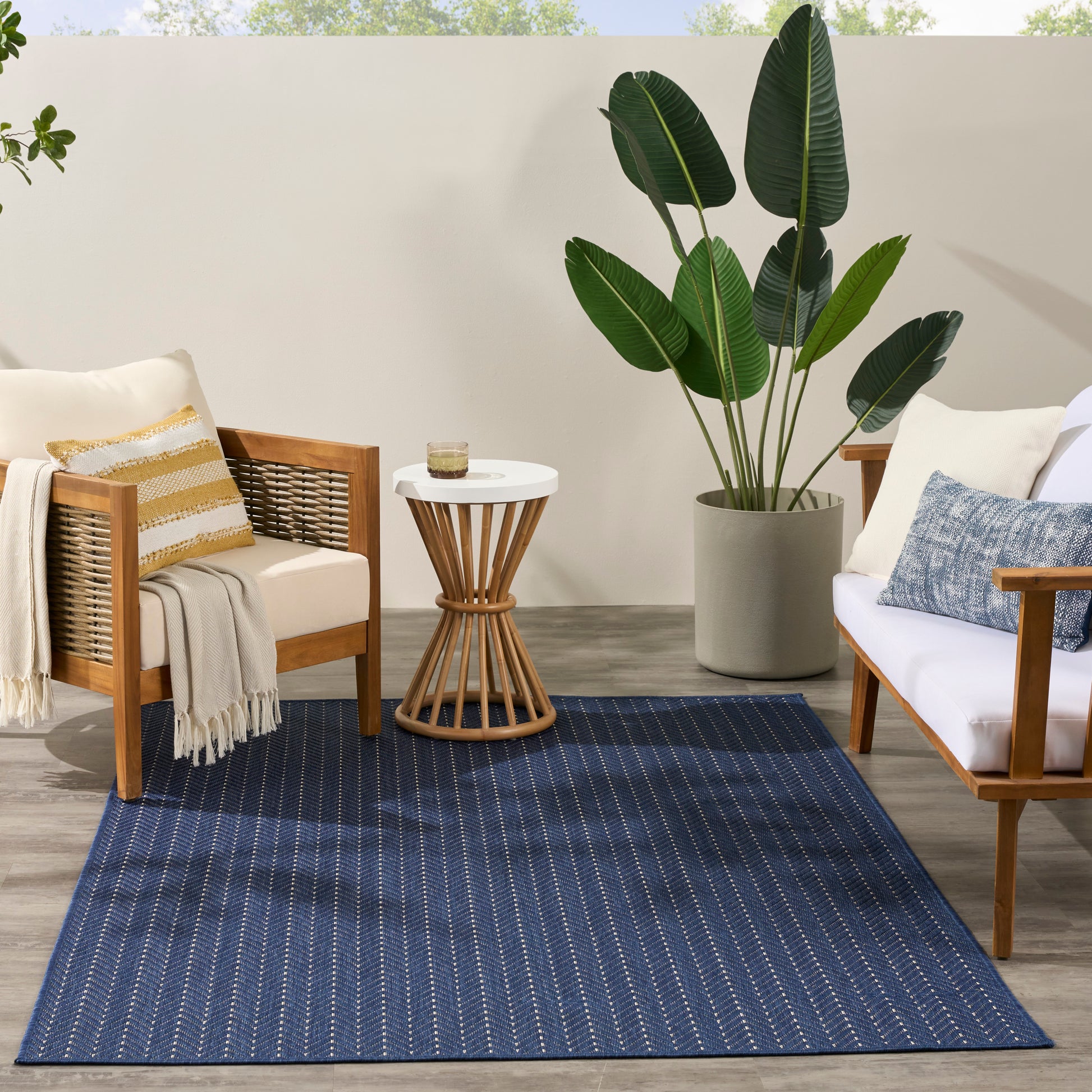 Blue woven outdoor rug with white dotted lines between wooden patio chairs, a round side table, and a tall potted plant.