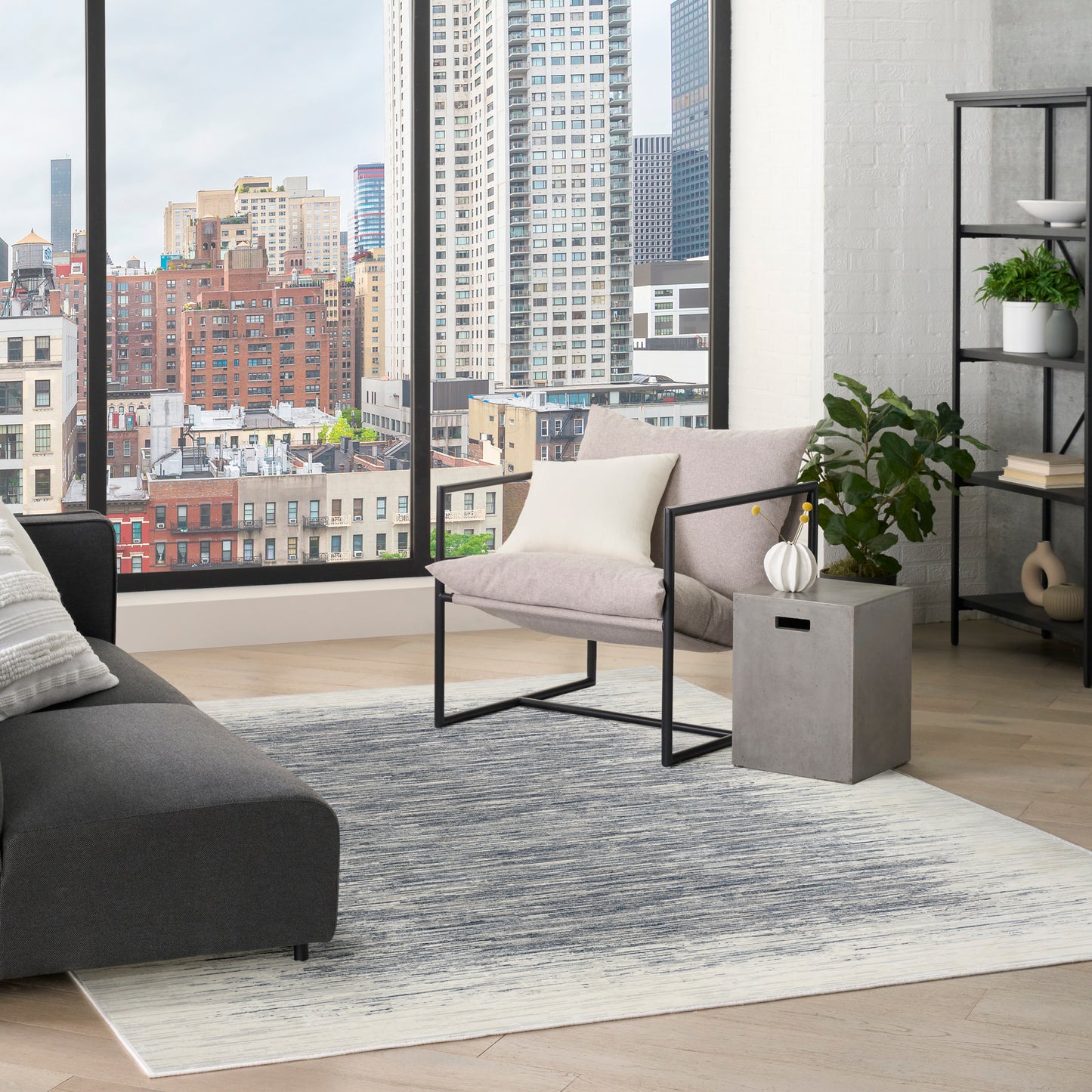 Modern living room with city view, gray sofa, beige armchair with black frame, concrete side table, plant, and gradient rug.