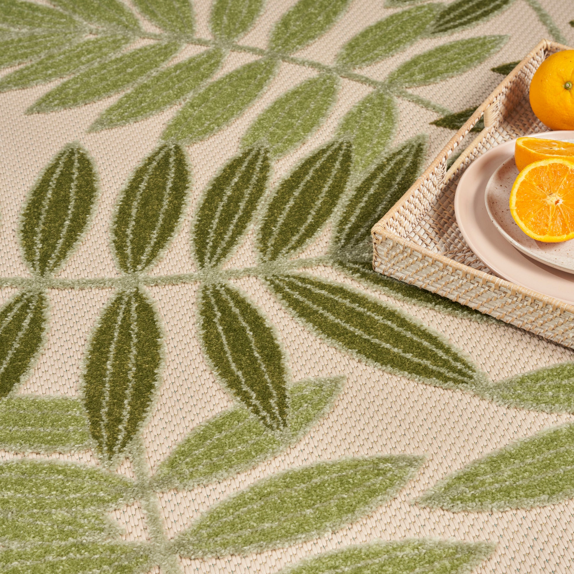 Beige woven rug featuring green leafy pattern; wicker tray with a plate and halved orange.