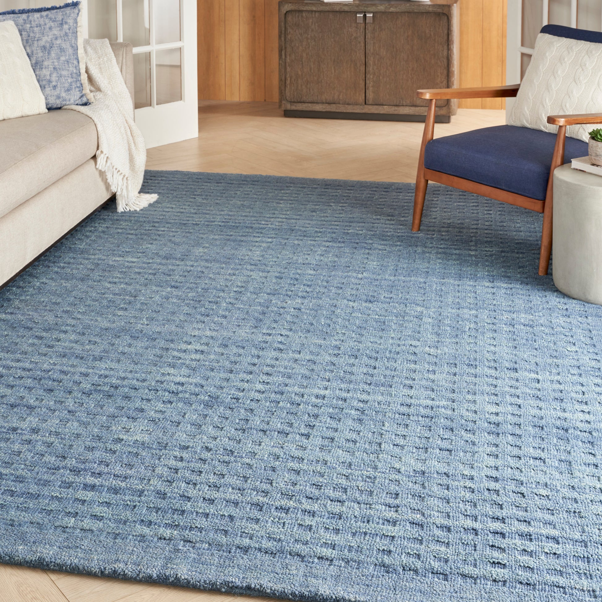 Blue textured area rug in a cozy living room with beige sofa, navy armchair, cream throw, and wooden cabinet.