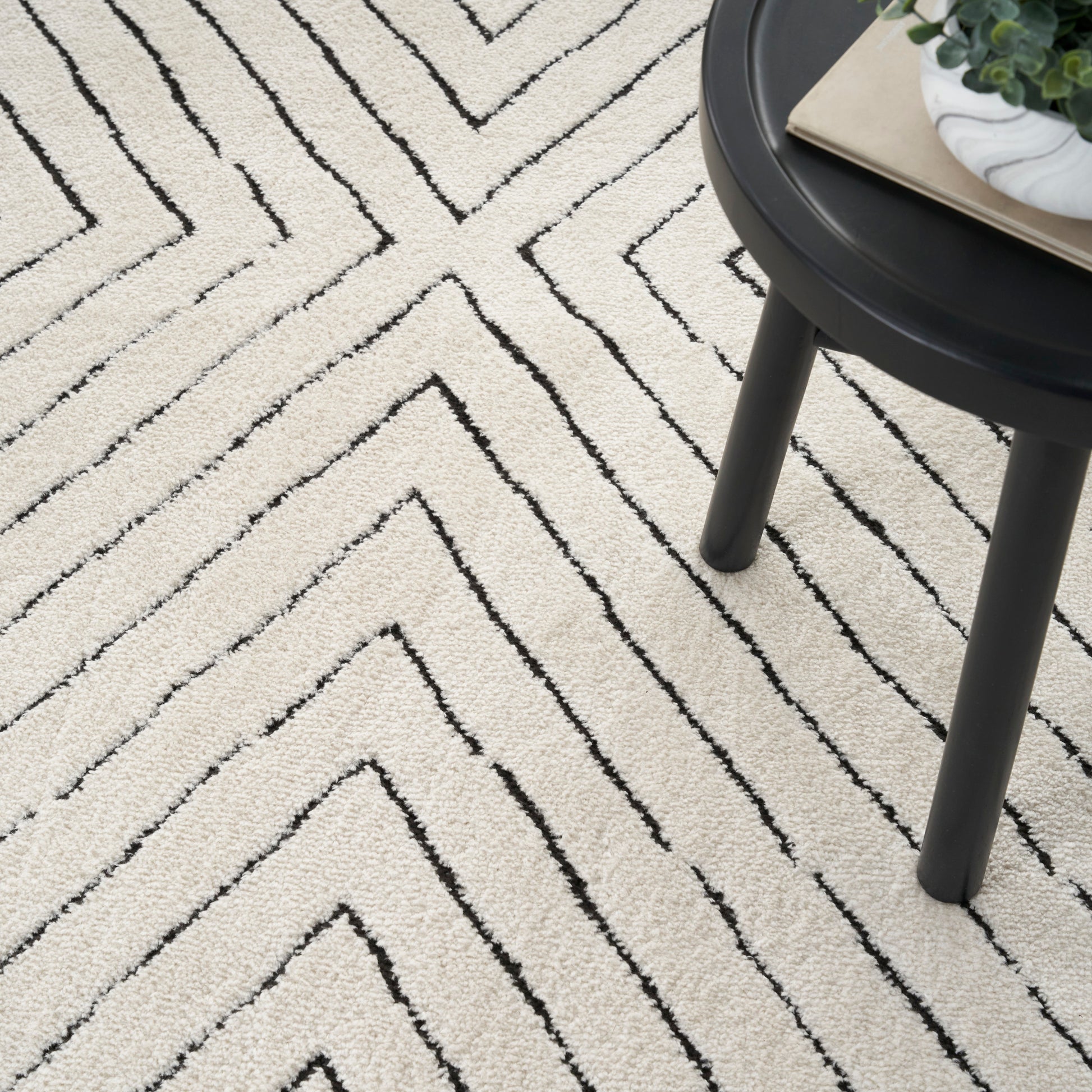 Beige rug with bold black chevron lines; round dark table topped with a plant and a book.