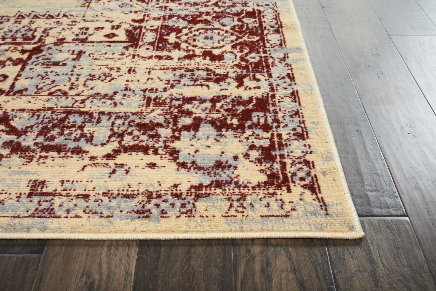 Distressed beige oriental rug with burgundy floral pattern and light blue accents, fringed edge on dark wood floor.