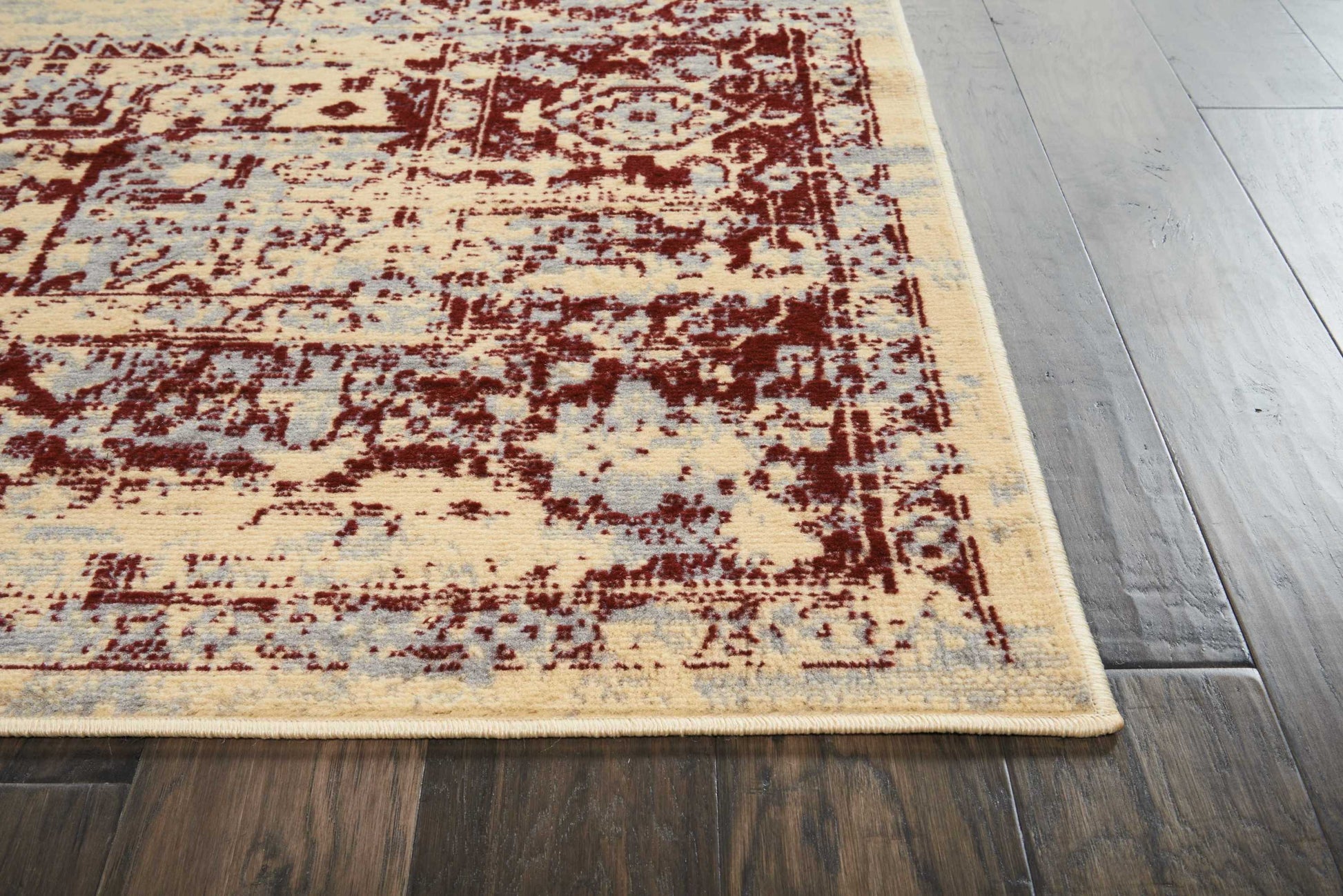 Distressed beige oriental rug with burgundy floral pattern and light blue accents, fringed edge on dark wood floor.