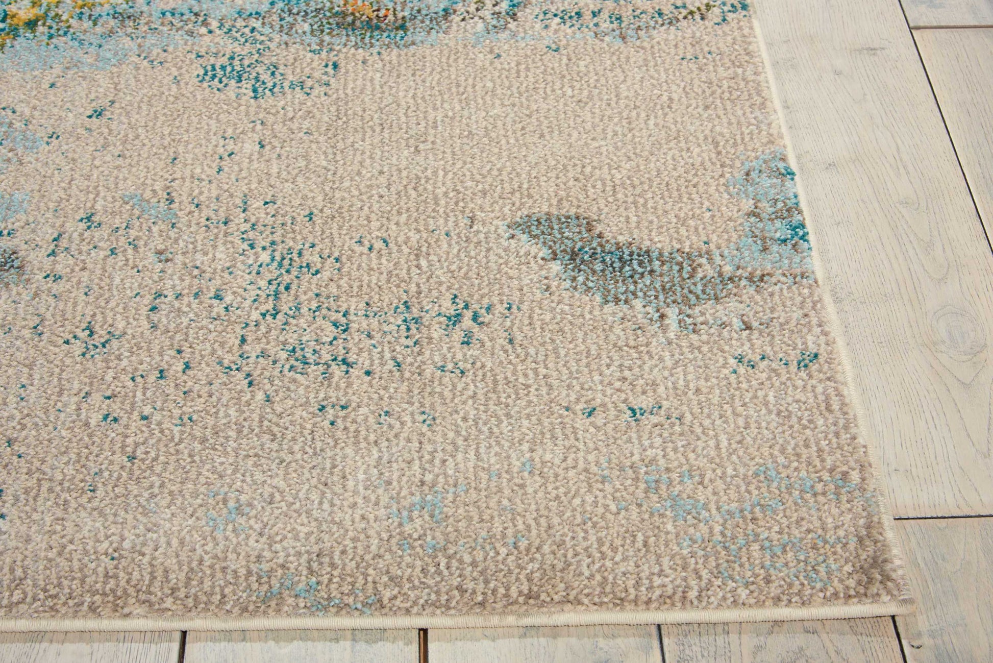 Beige rectangular flat-weave rug with turquoise and teal abstract pattern on light wooden floor.