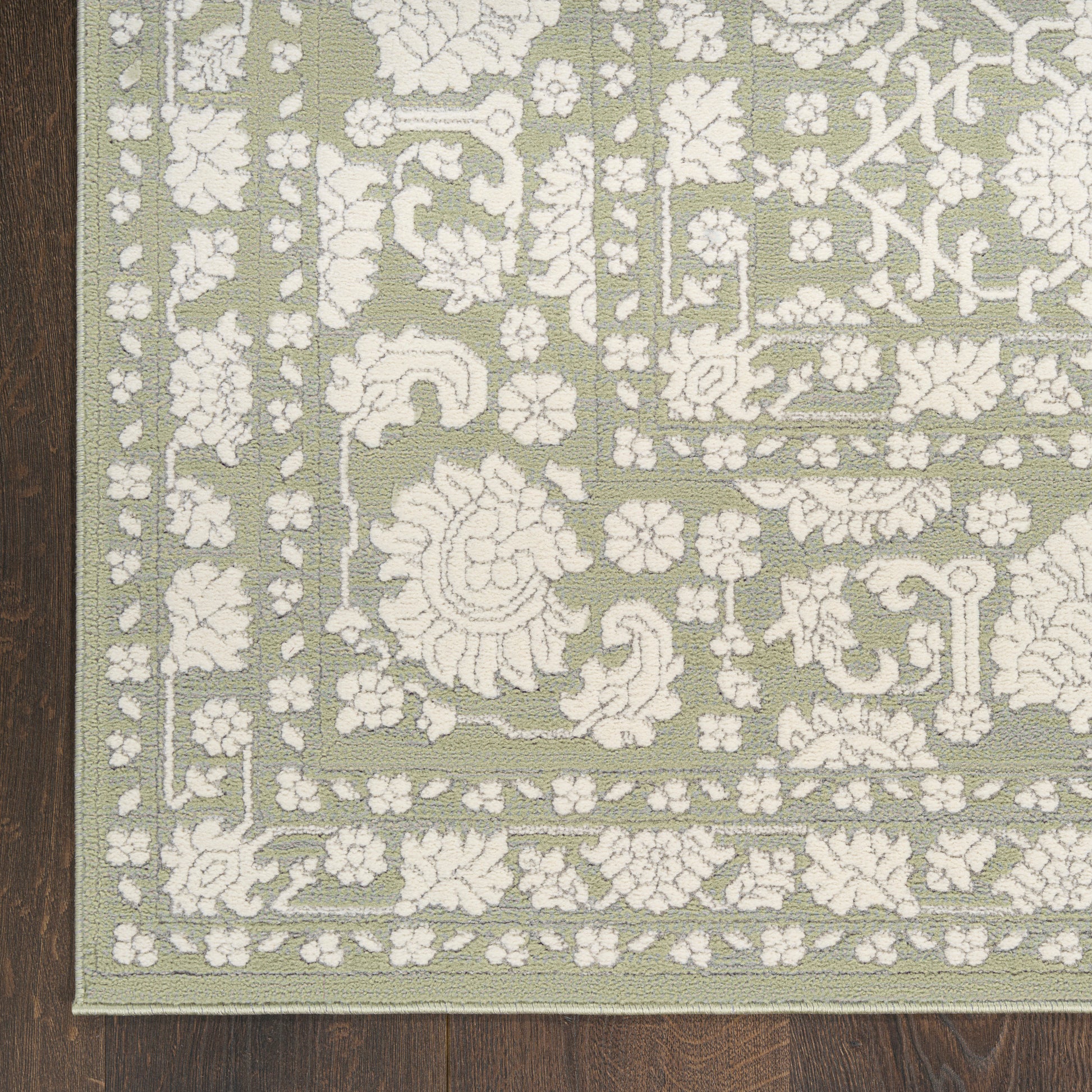 Sage green rug with white floral damask pattern and decorative border on dark wooden floor.