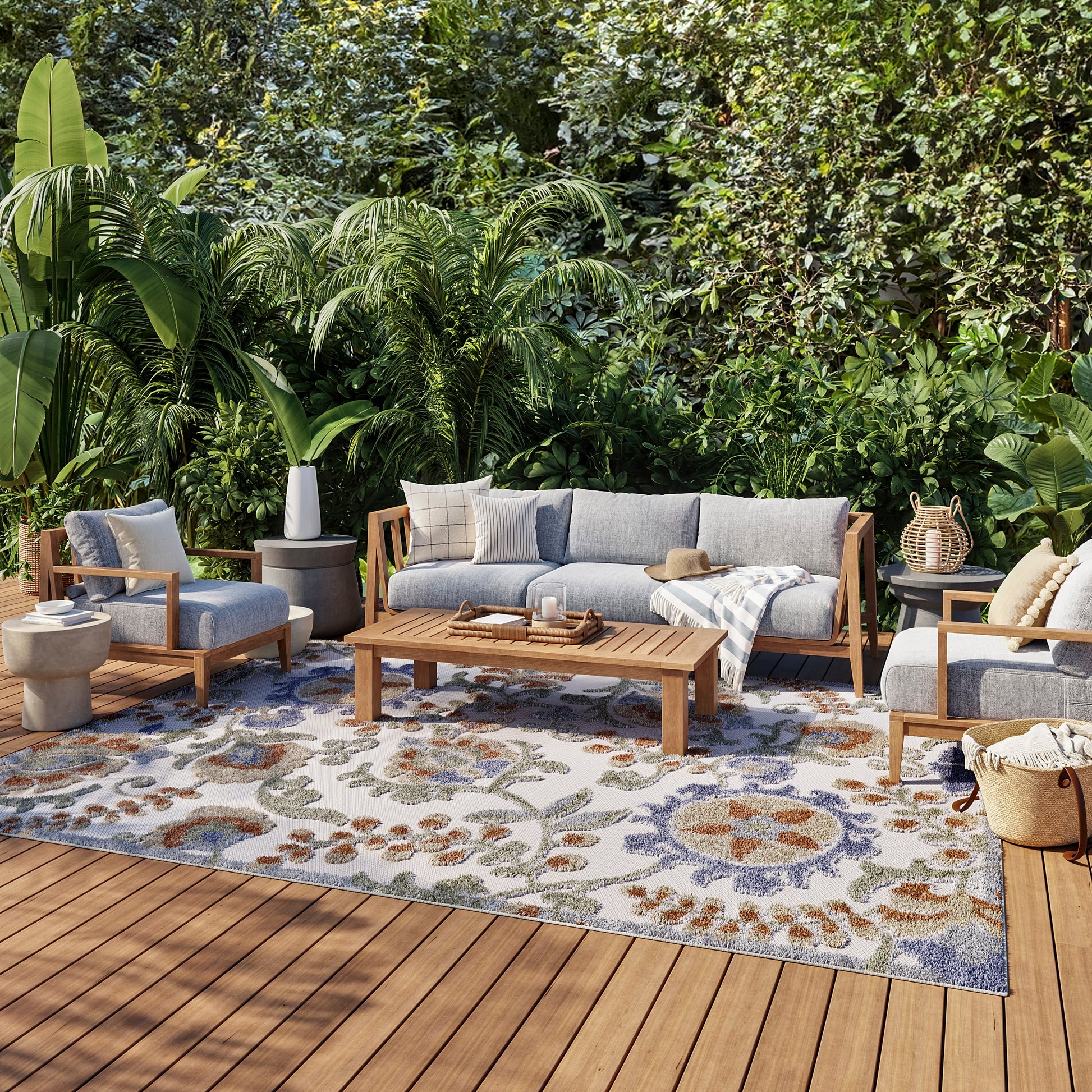 Outdoor lounge set with blue-gray cushions on wooden frames, coffee table, and a floral rug amid tropical plants.