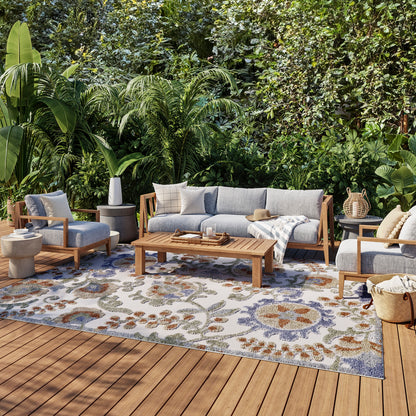 Outdoor lounge set with blue-gray cushions on wooden frames, coffee table, and a floral rug amid tropical plants.