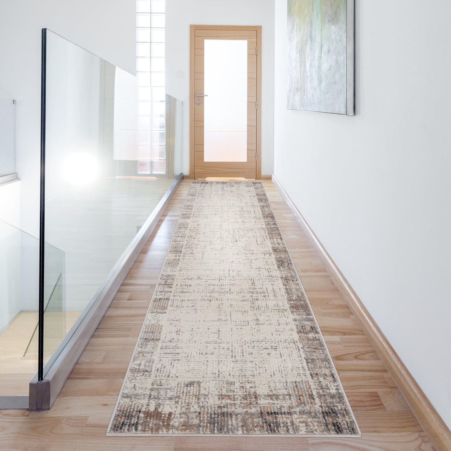 Beige patterned hallway runner rug on light wood floor, glass railing on the left, wooden door at the end, abstract wall art on the right.