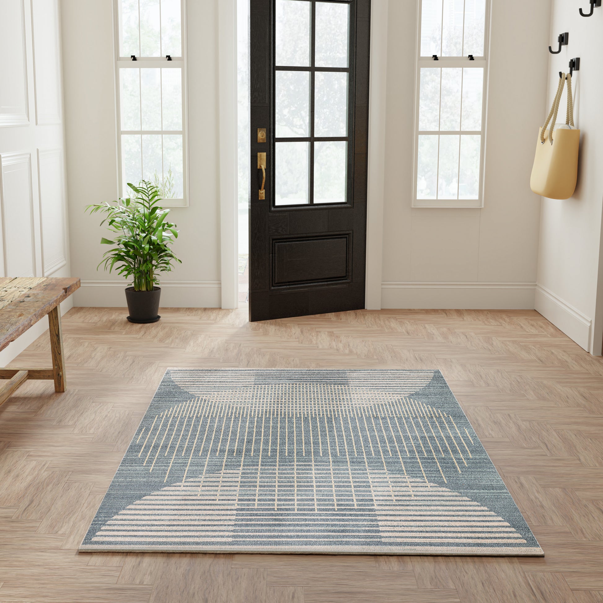 Blue geometric woven area rug in entryway with black front door, light wood floor, plant, bench, and tote bag.