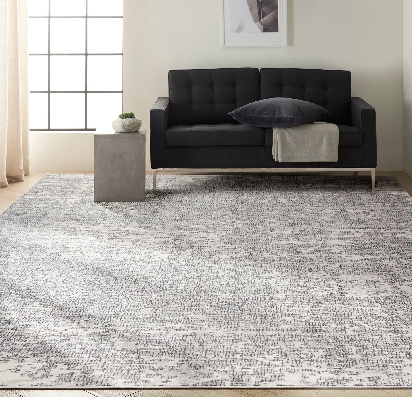 Dark gray tufted loveseat with metal legs, beige throw, charcoal pillow on a light gray rug; concrete side table with plant.