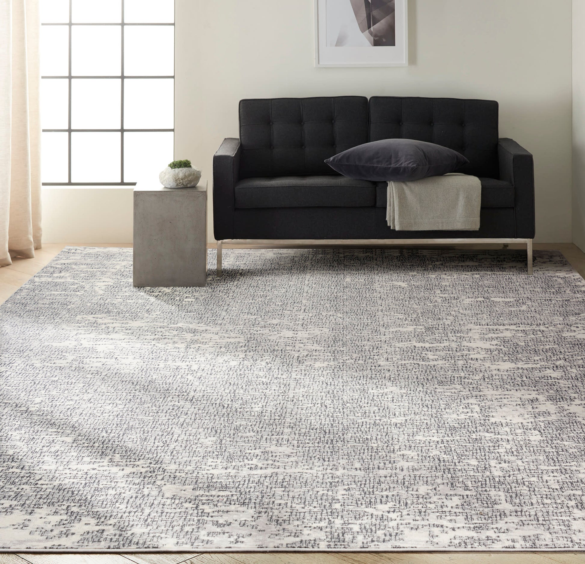 Dark gray tufted loveseat with metal legs, beige throw, charcoal pillow on a light gray rug; concrete side table with plant.