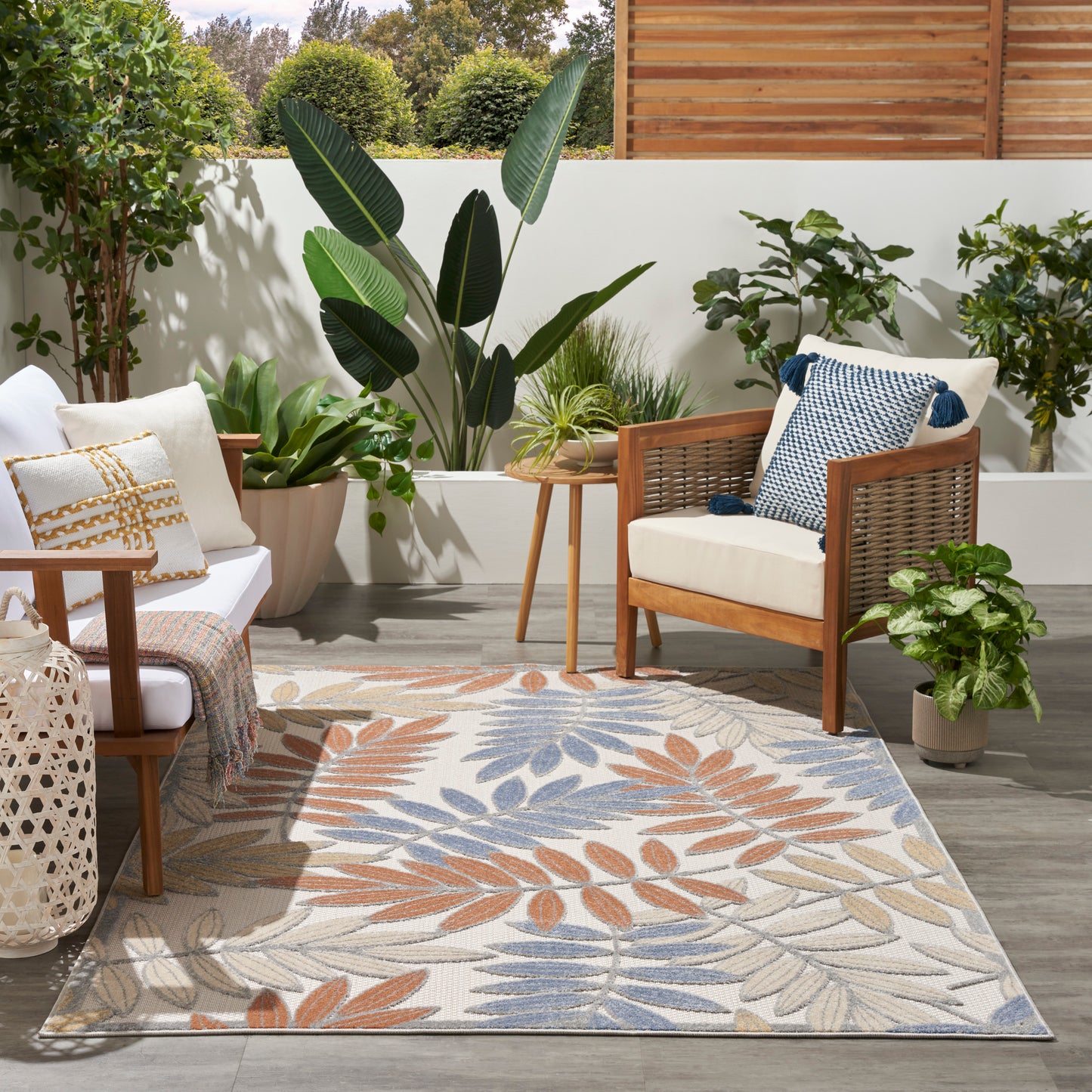 Outdoor seating with wooden-framed chairs, beige cushions, a leaf-pattern rug, side table, and lush potted plants.