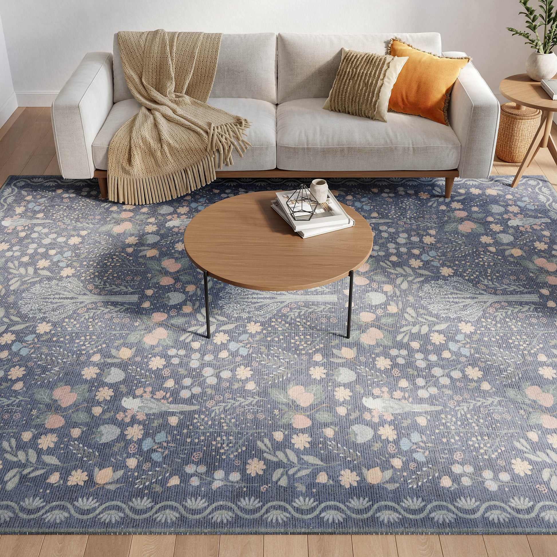 Light gray sofa with beige throw blanket, orange and striped cushions; round wooden coffee table on a blue floral area rug.