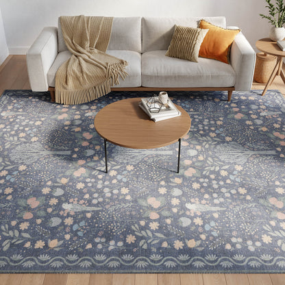Light gray sofa with beige throw blanket, orange and striped cushions; round wooden coffee table on a blue floral area rug.