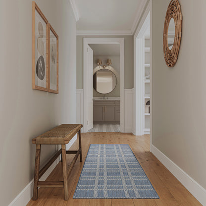 Neutral hallway with gray walls and wainscoting, wooden floor, a blue runner rug, bench, framed art, wicker round mirror, leading to the bathroom.