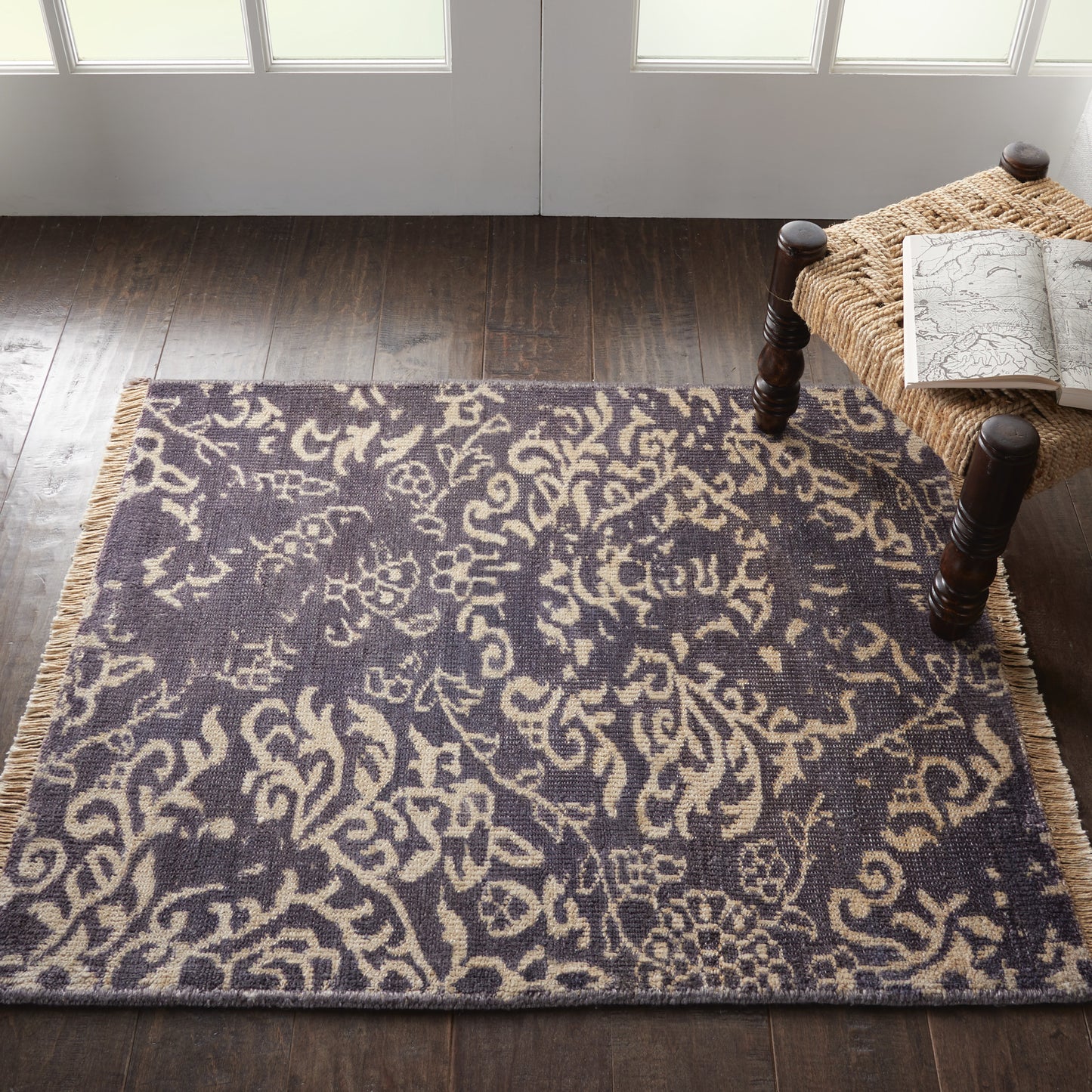 Dark blue area rug with beige damask pattern and fringe on a wood floor, beside a wicker bench with a map.