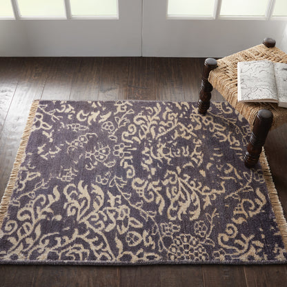 Dark blue area rug with beige damask pattern and fringe on a wood floor, beside a wicker bench with a map.