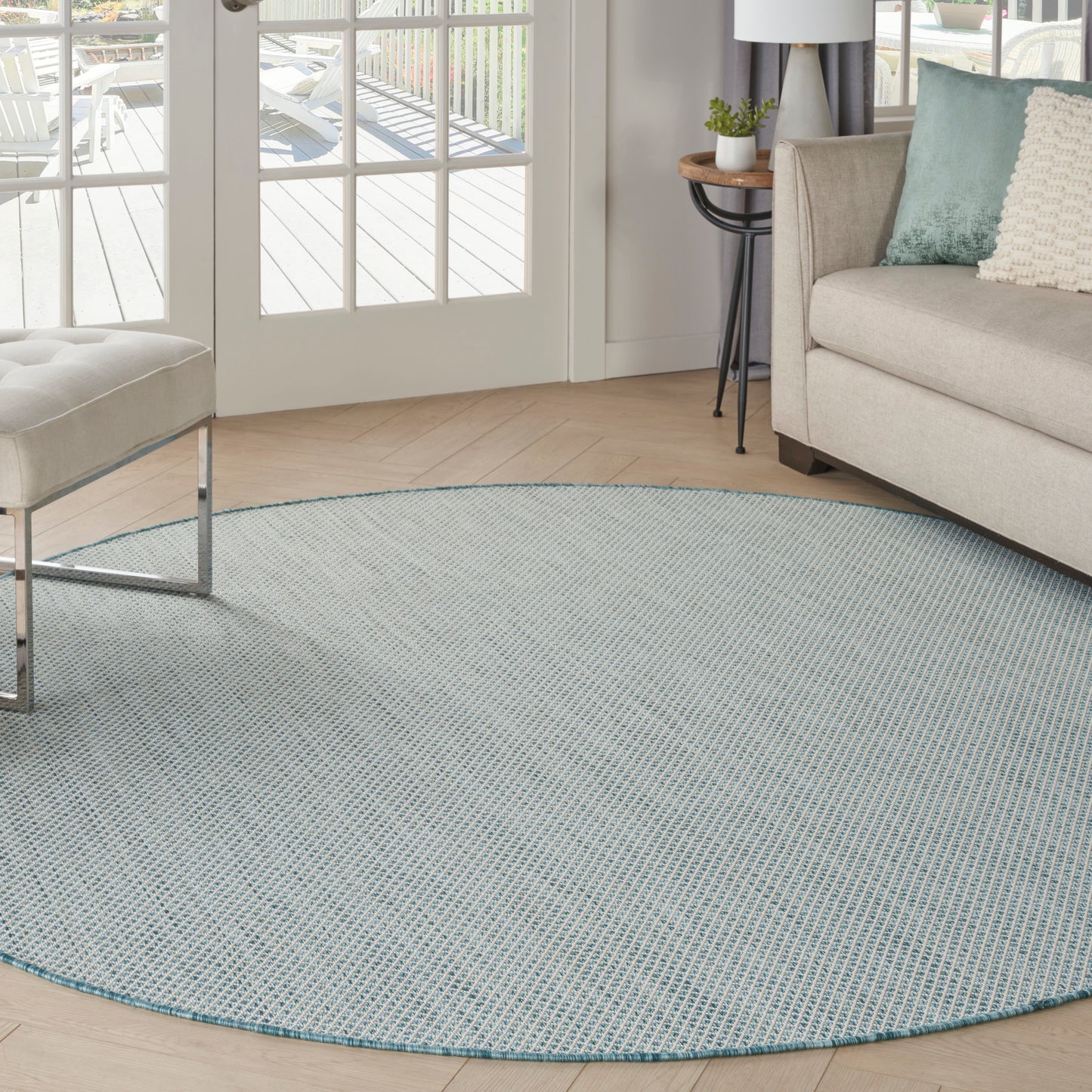 Round blue-gray woven round area rug on light wood floor in a modern living room with a beige sofa and teal cushions.