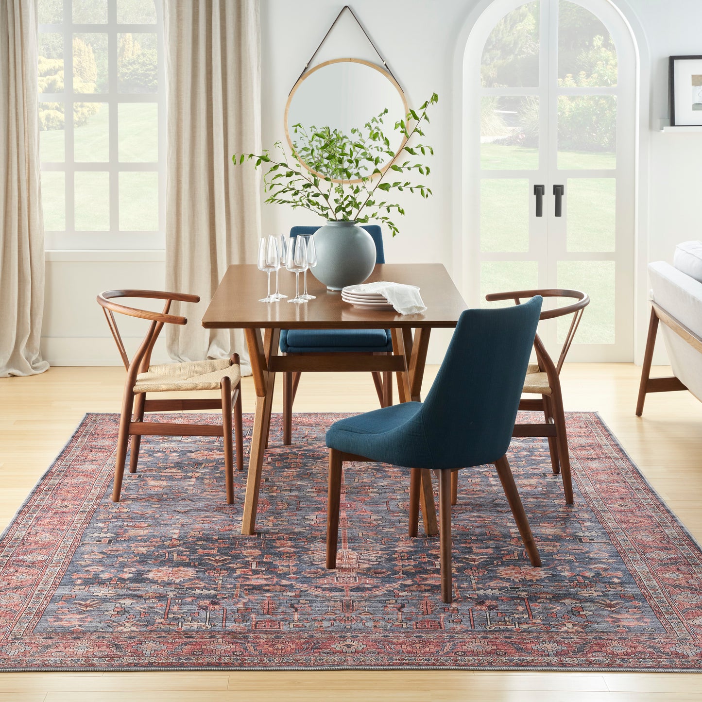 Sunlit dining nook with wood table, blue upholstered chair, wooden chairs, rug, greenery vase, and round gold mirror.