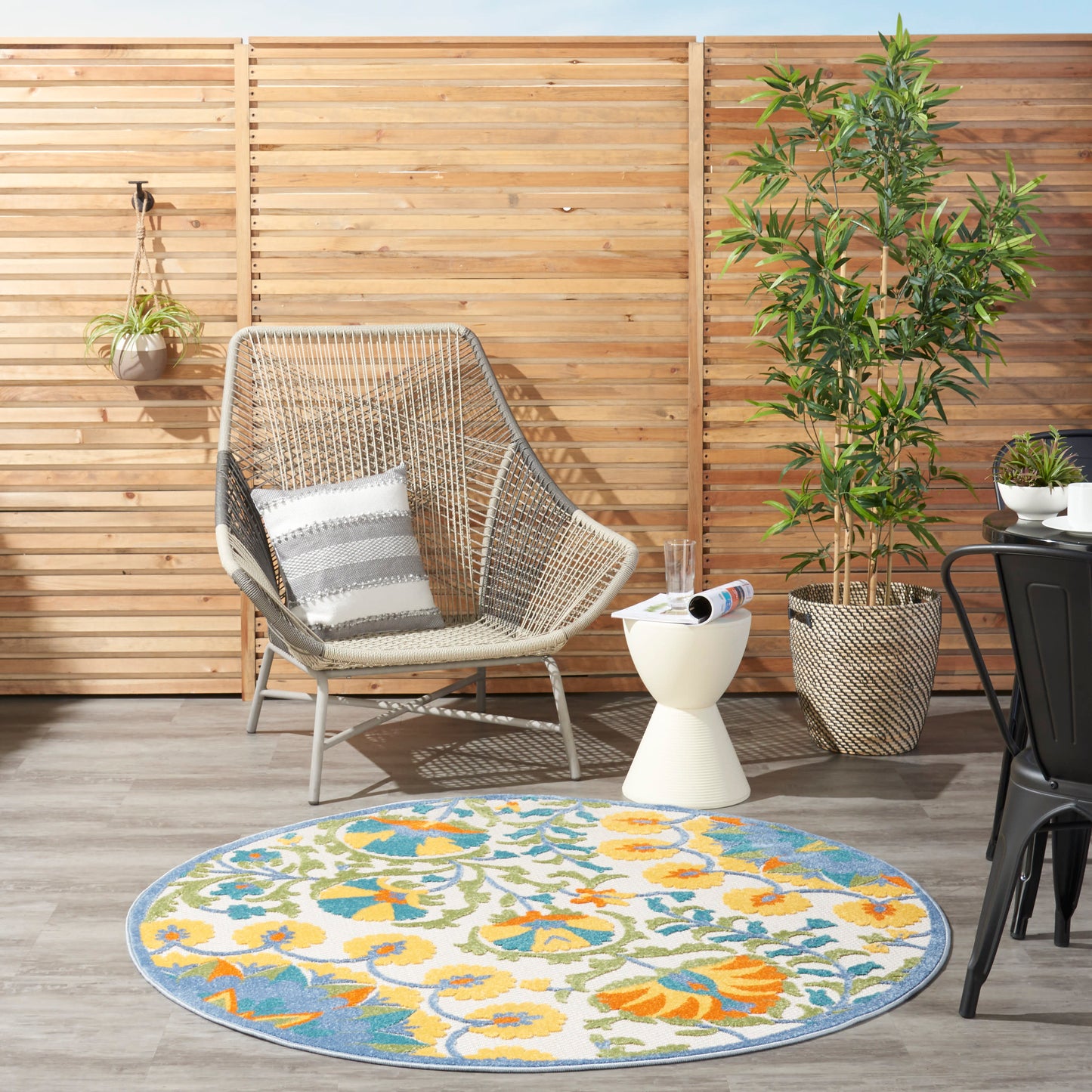 Outdoor patio with wicker chair, striped cushion, white side table, round floral blue-yellow rug, and potted plants.