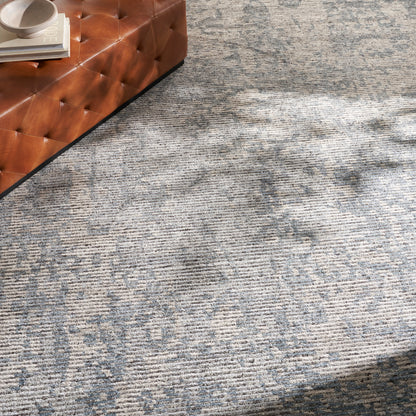Blue-gray patterned rug with a brown tufted leather ottoman in the corner and a white cup atop magazines.