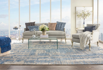 Bright modern living room with beige sofa, blue patterned cushions, glass coffee table, blue area rug, and plush armchairs.