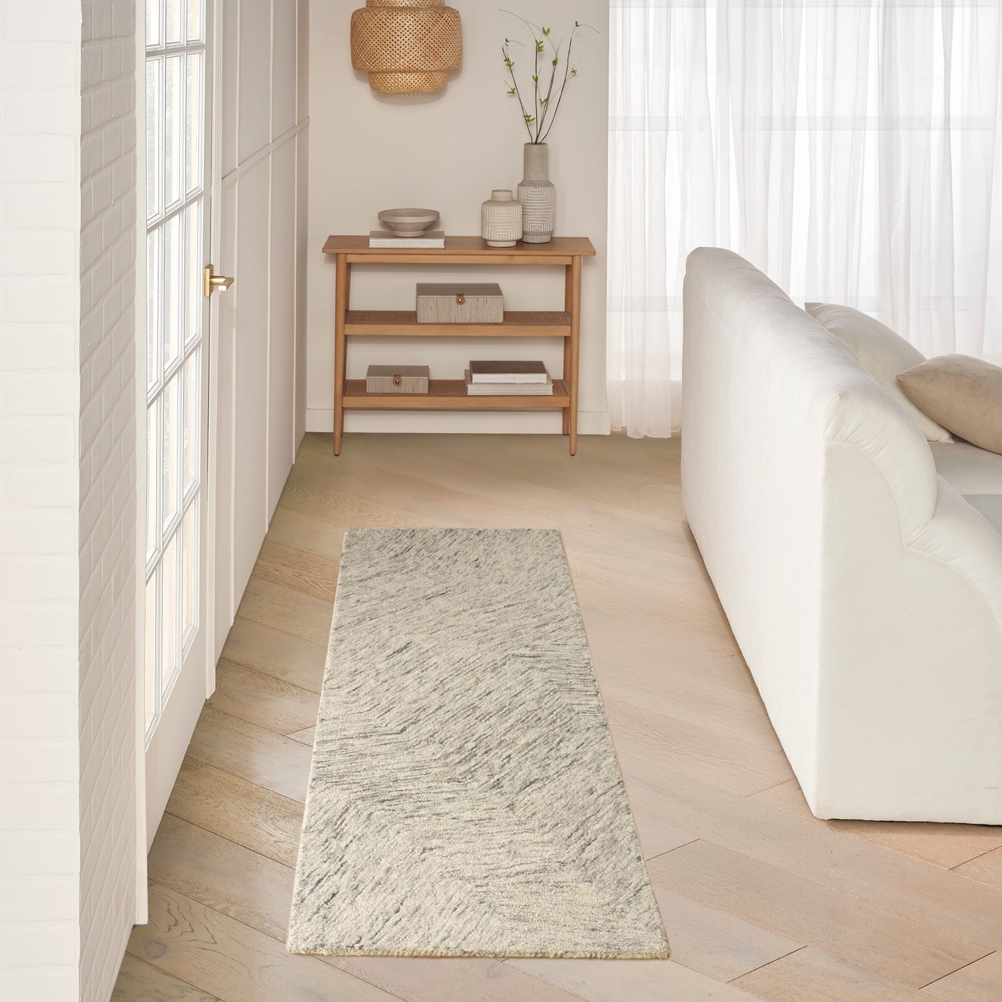 Minimalist light-wood hallway console with shelves, gray textured runner rug, white sofa and sheer curtains.