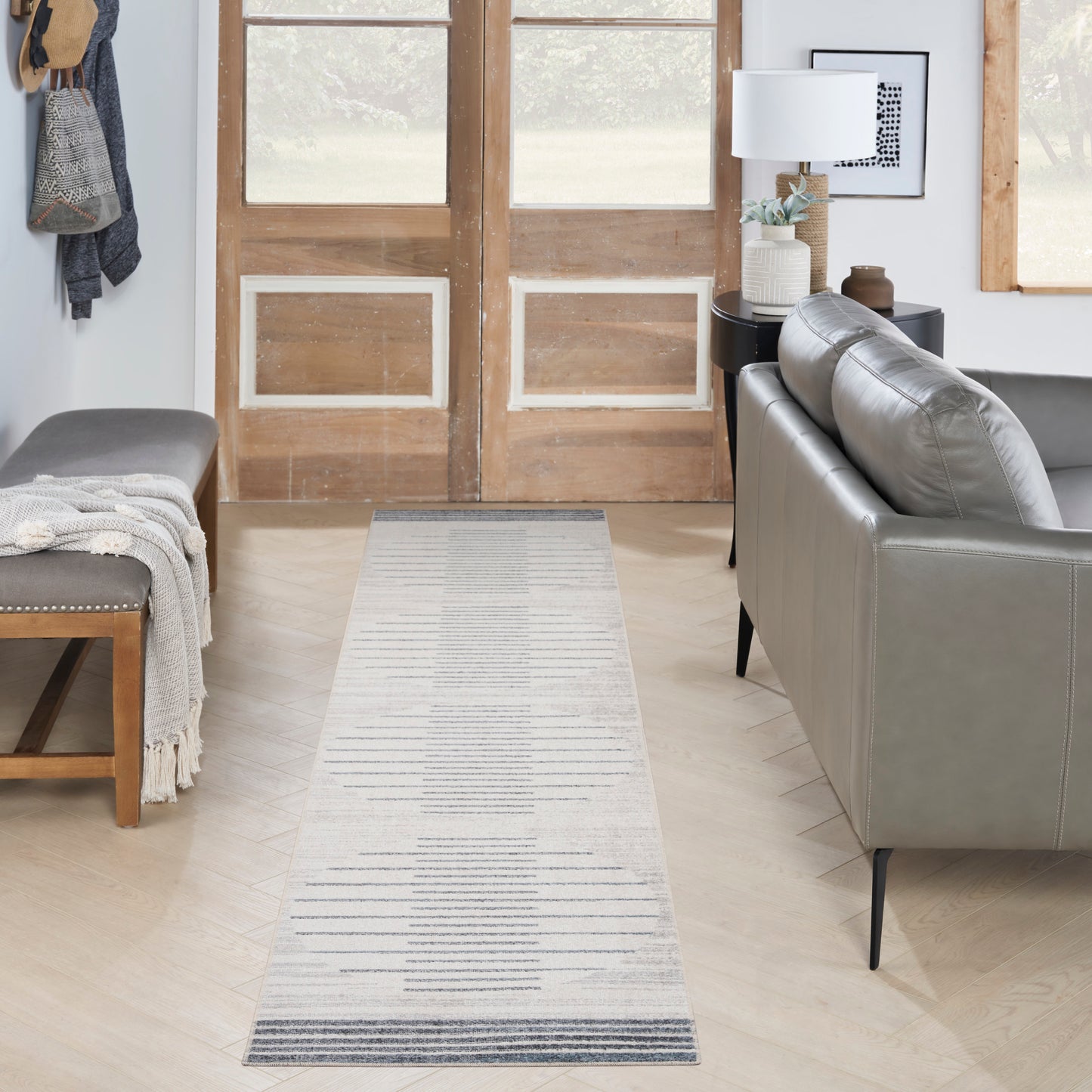 Light gray striped runner rug on light wood floor, in a modern living space with a gray leather sofa, bench, and wooden doors.