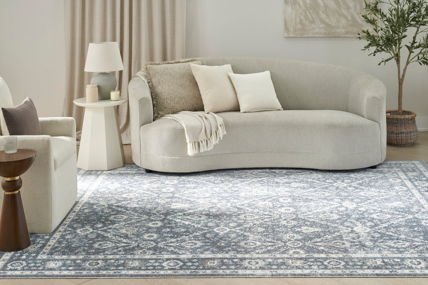 Light gray curved fabric sofa with cream cushions and a throw, set in a cozy modern living room with a blue ornate rug.