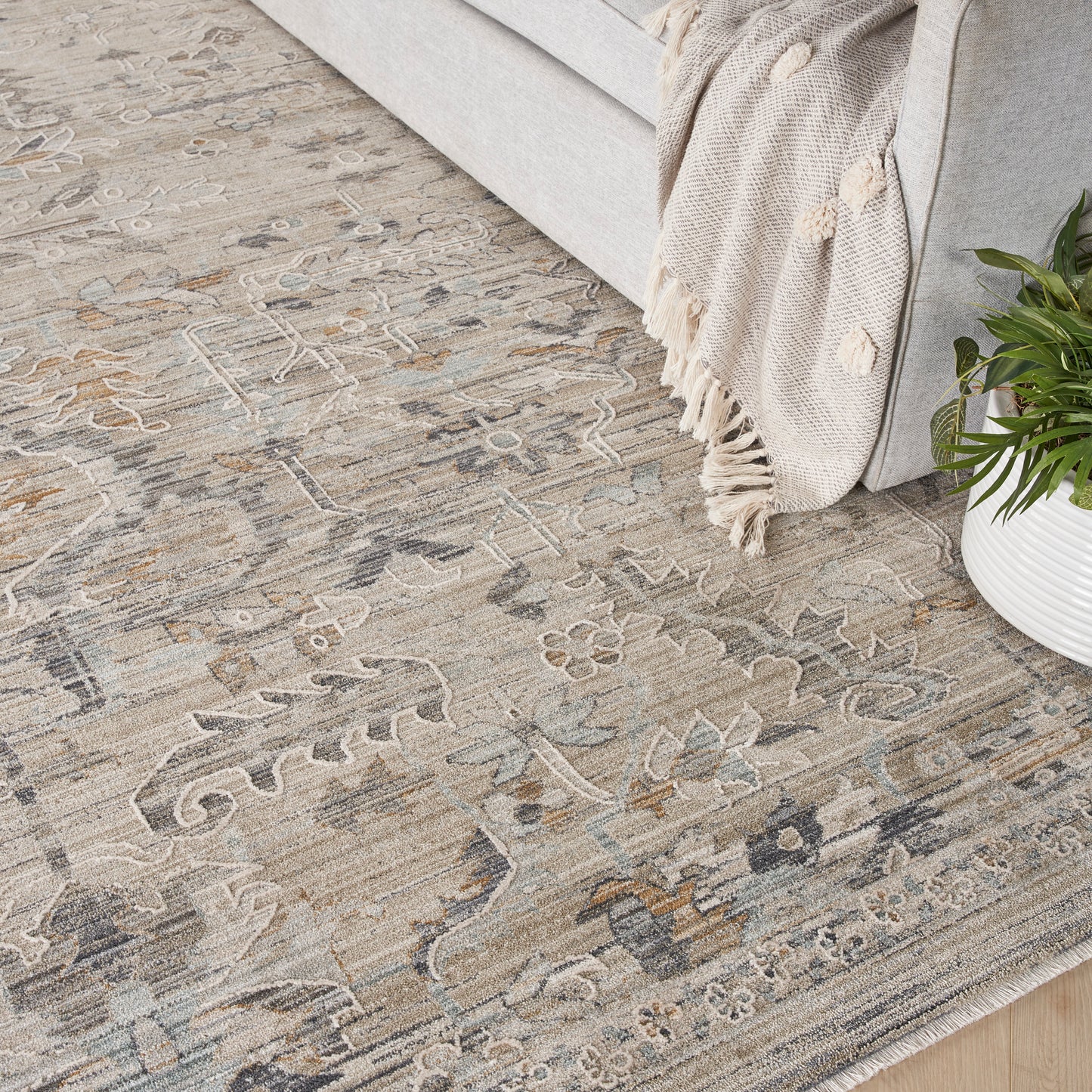 Muted beige-gray oriental rug with floral geometric pattern under a light gray sofa; fringed throw and potted plant nearby.