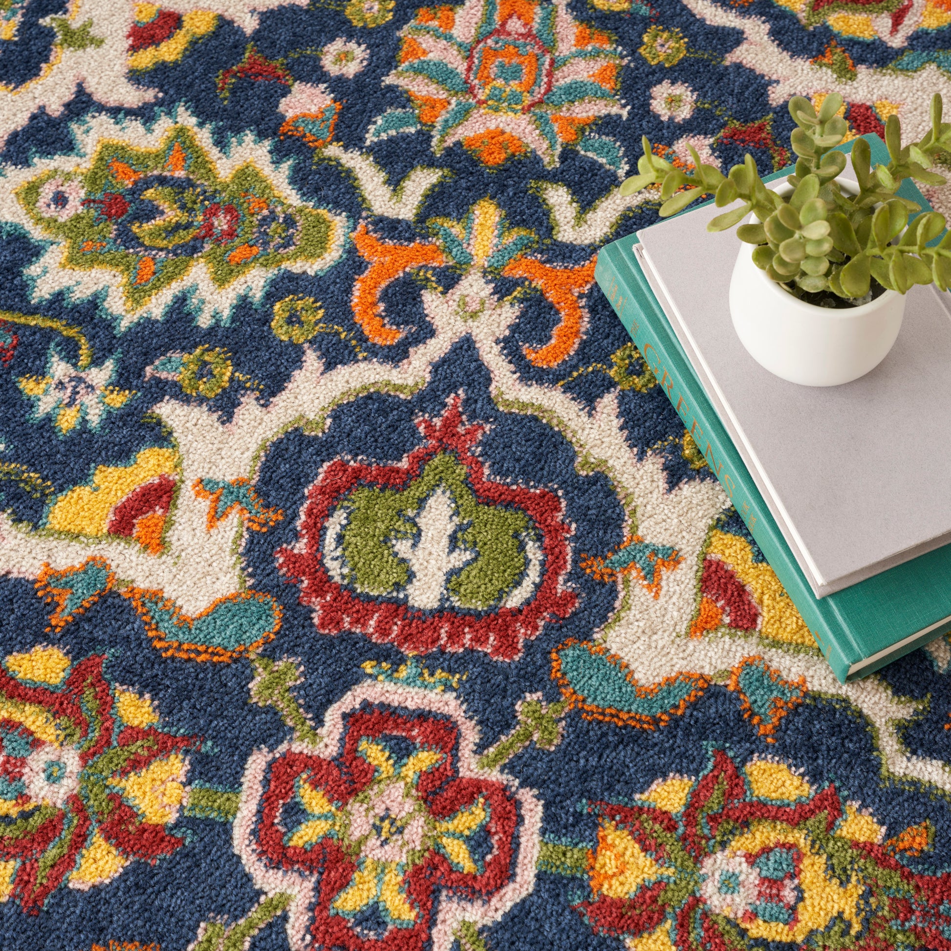 Vibrant navy rug with bold floral patterns in red, yellow, teal, and green; stack of books with a small potted succulent.