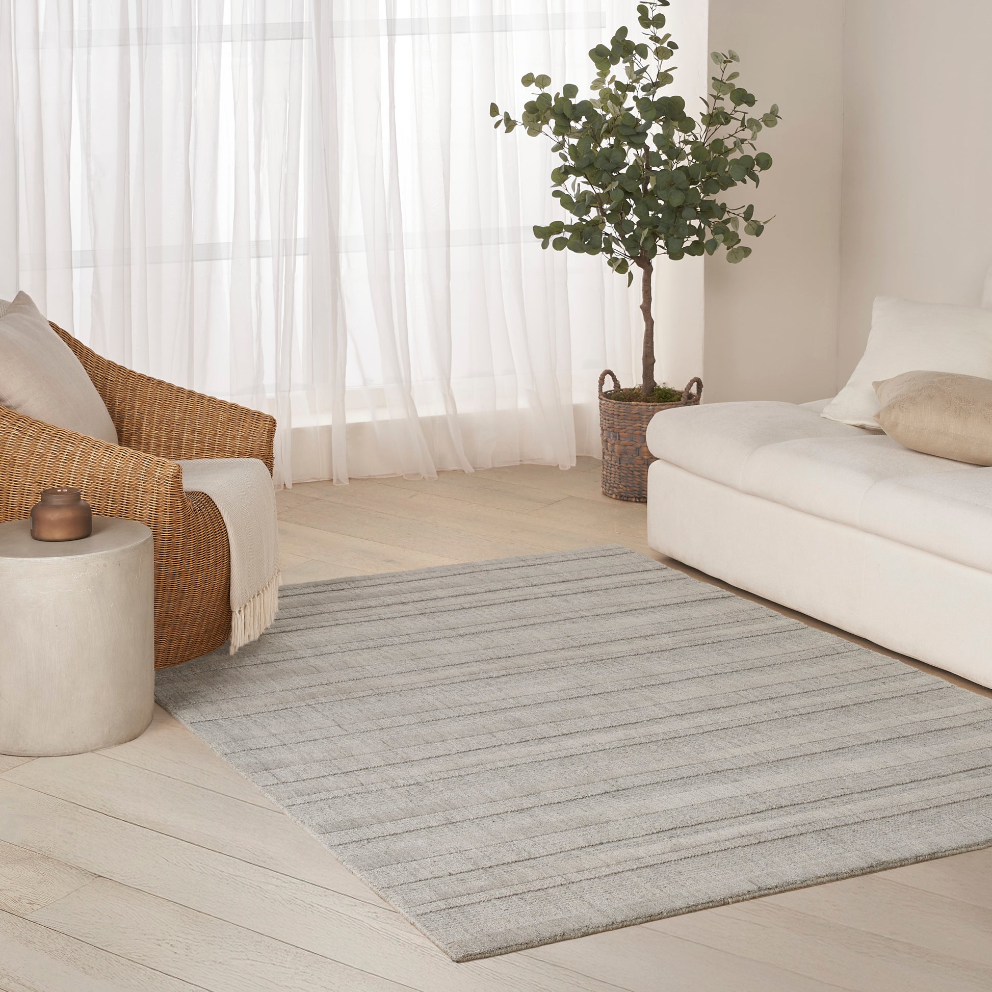 Bright living room with cream sofa, woven rattan chair, grey striped rug, potted plant in basket, sheer white curtains.