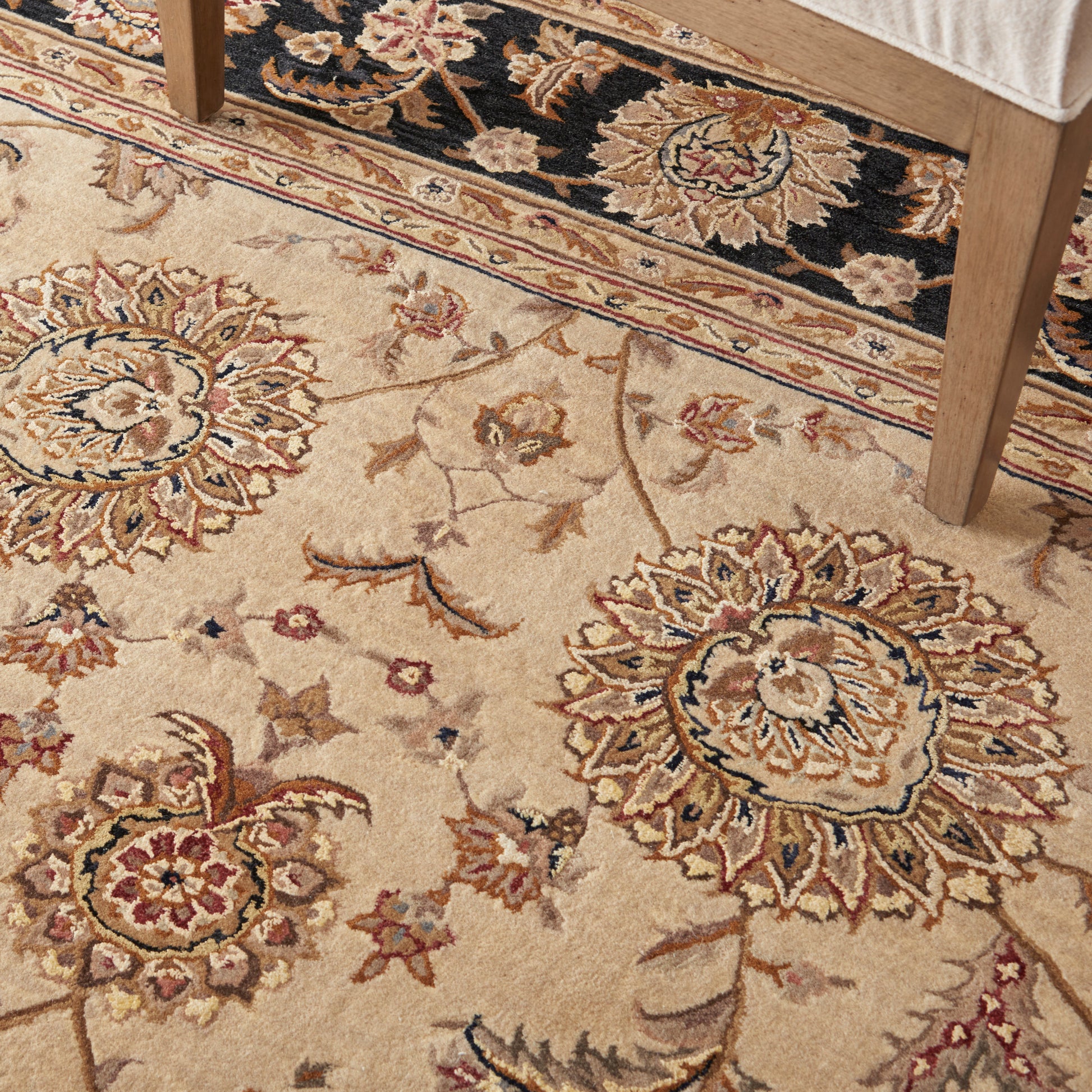 Beige traditional floral area rug with large medallions and blue accents; chair leg visible.