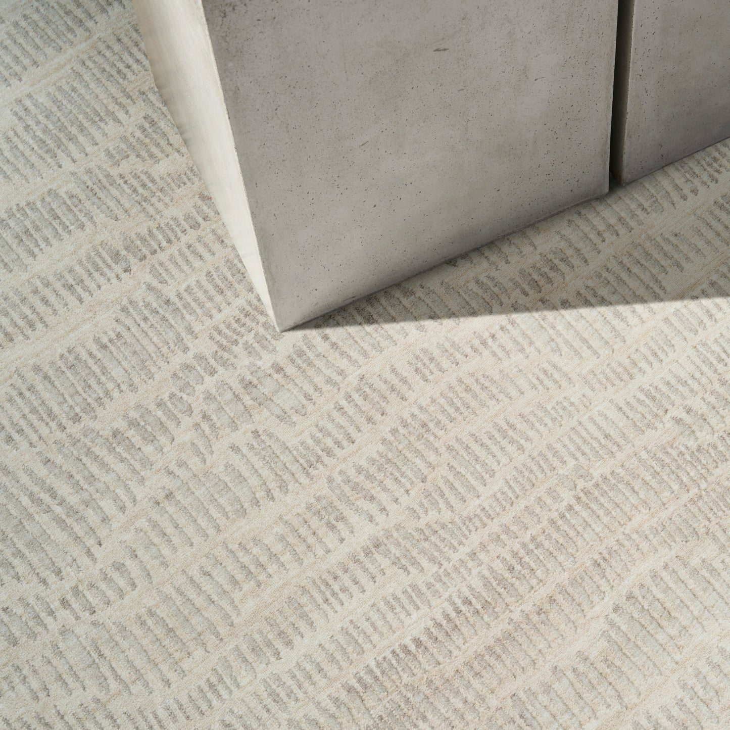 Beige textured rug with a subtle geometric weave, shown near light gray concrete blocks.