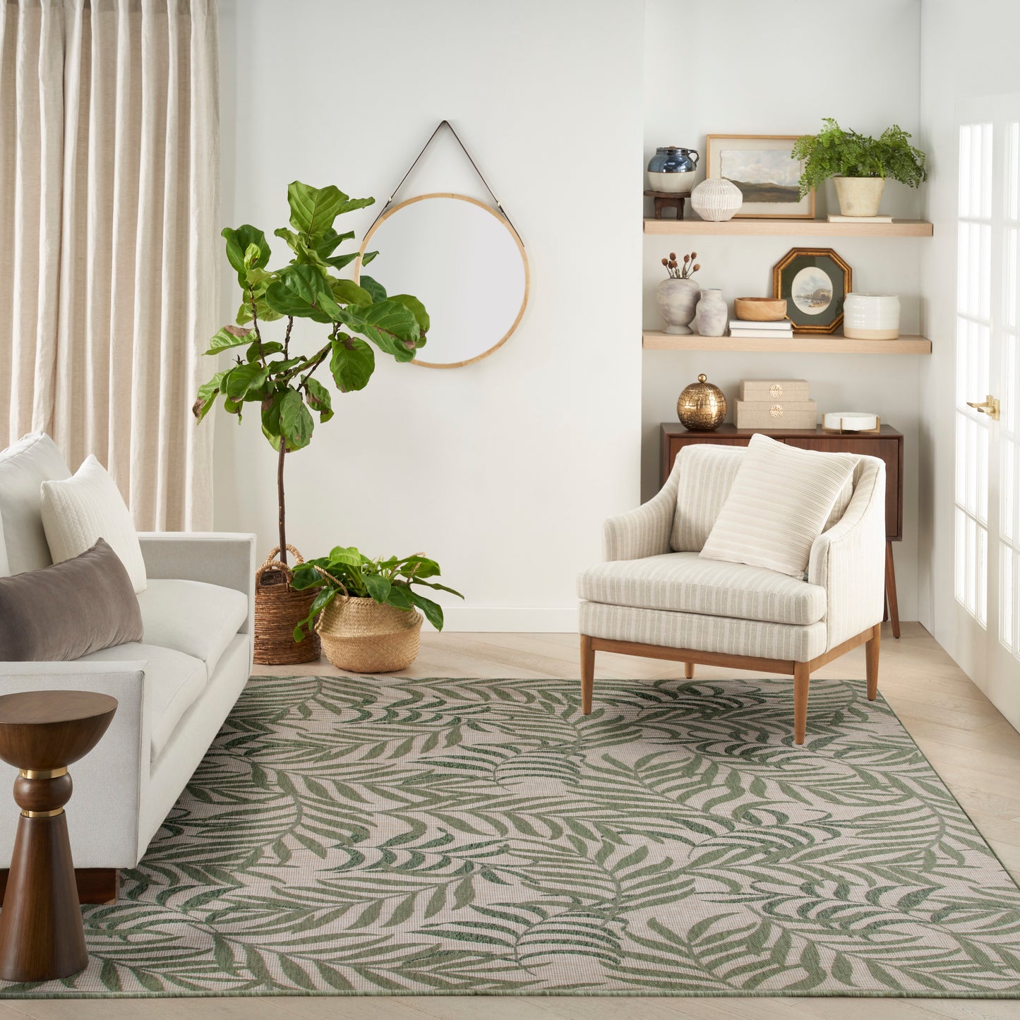 Neutral living room with cream sofa, striped armchair, palm-leaf rug, fiddle-leaf fig plants, round wall mirror, shelves.