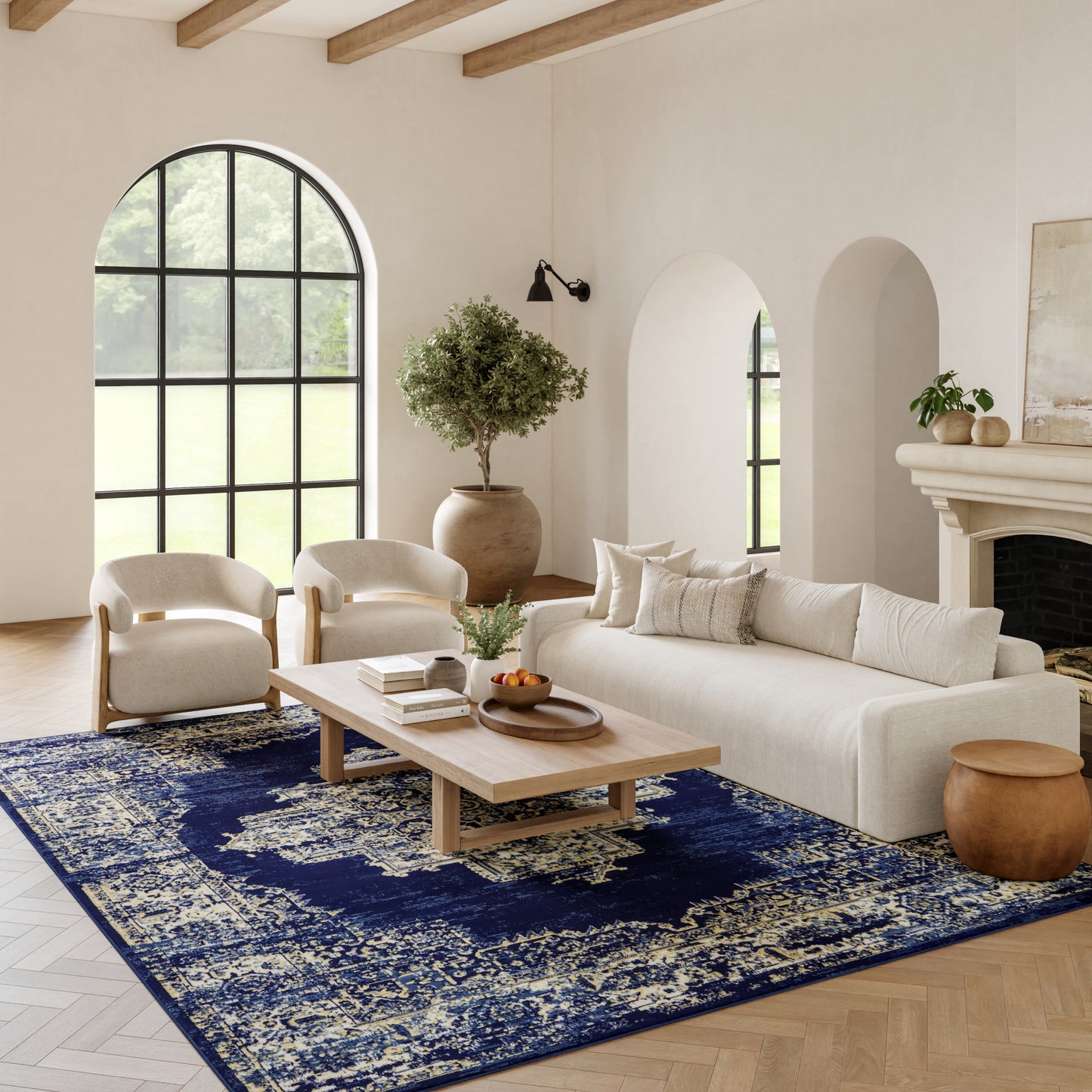 Neutral living room with arched window, cream sofa and chairs, light wood coffee table, navy distressed rug, potted tree.