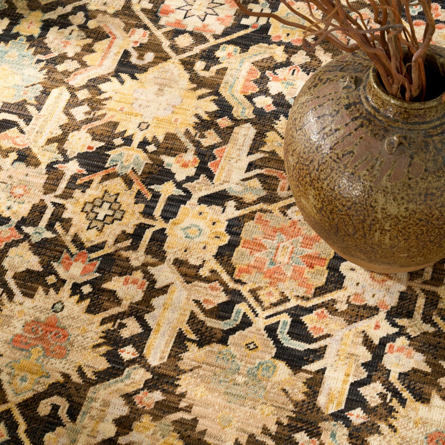 Oriental rug with cream, tan, black geometric motif and pale blue accents; brown ceramic vase with dried branches.