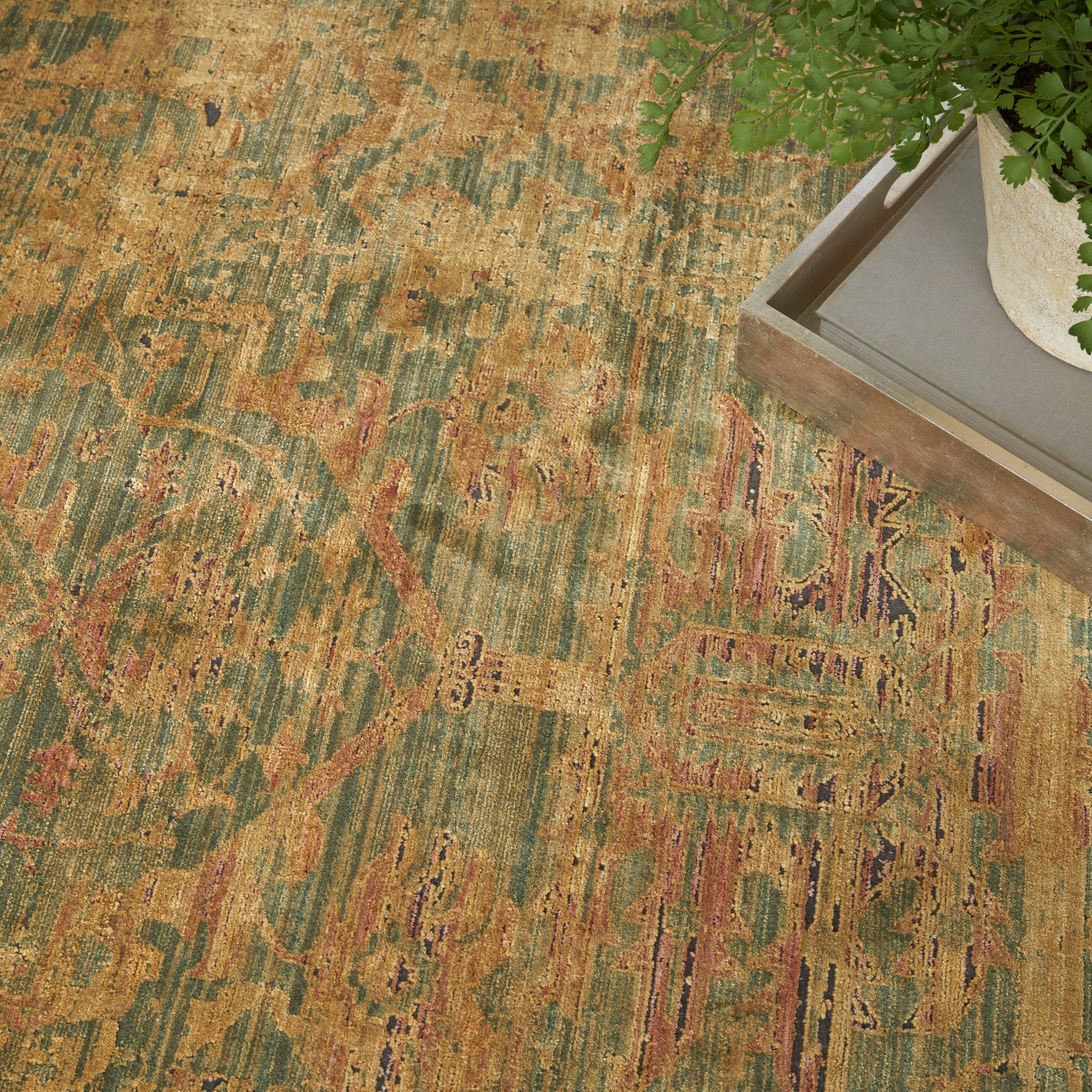 Distressed oriental rug in green, gold and rust with faded medallion pattern; wooden tray with a potted plant in corner.