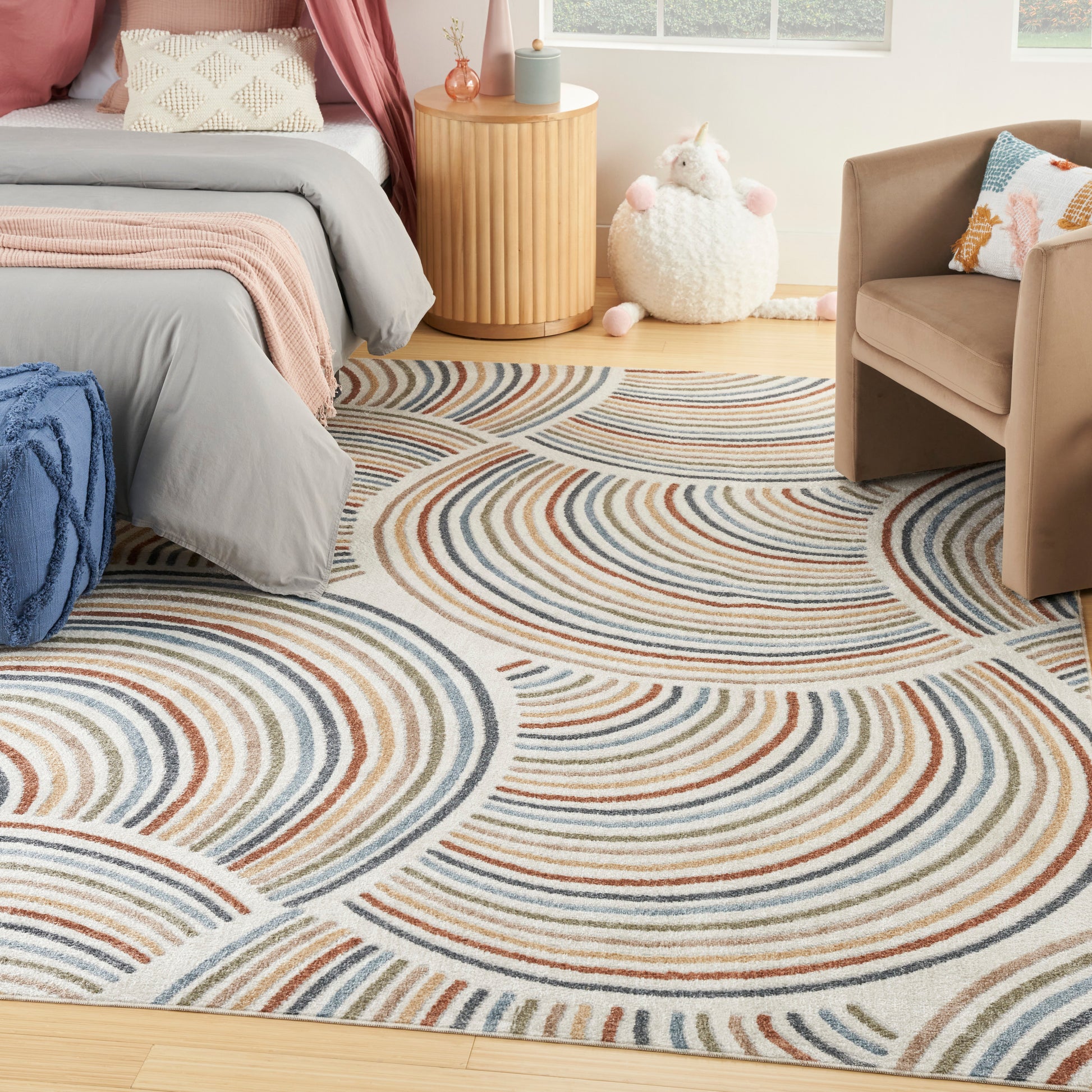 Cozy bedroom with a multicolor swirl rug, gray bedding with pink throw, beige chair, and a wooden side table.