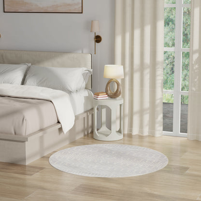 Cream upholstered bed with white bedding, white nightstand, lamp, books, gray rug, light wood floor, sheer curtains.