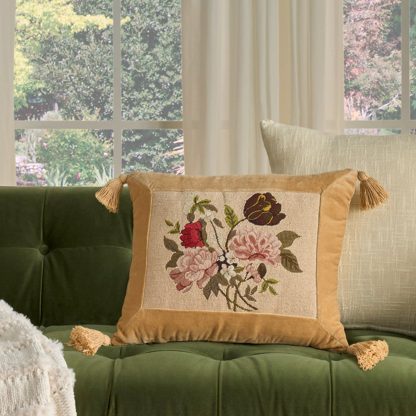 Beige square pillow 16x20 with gold satin-edged trim, tassels and pink/red floral embroidery on a tufted sofa.