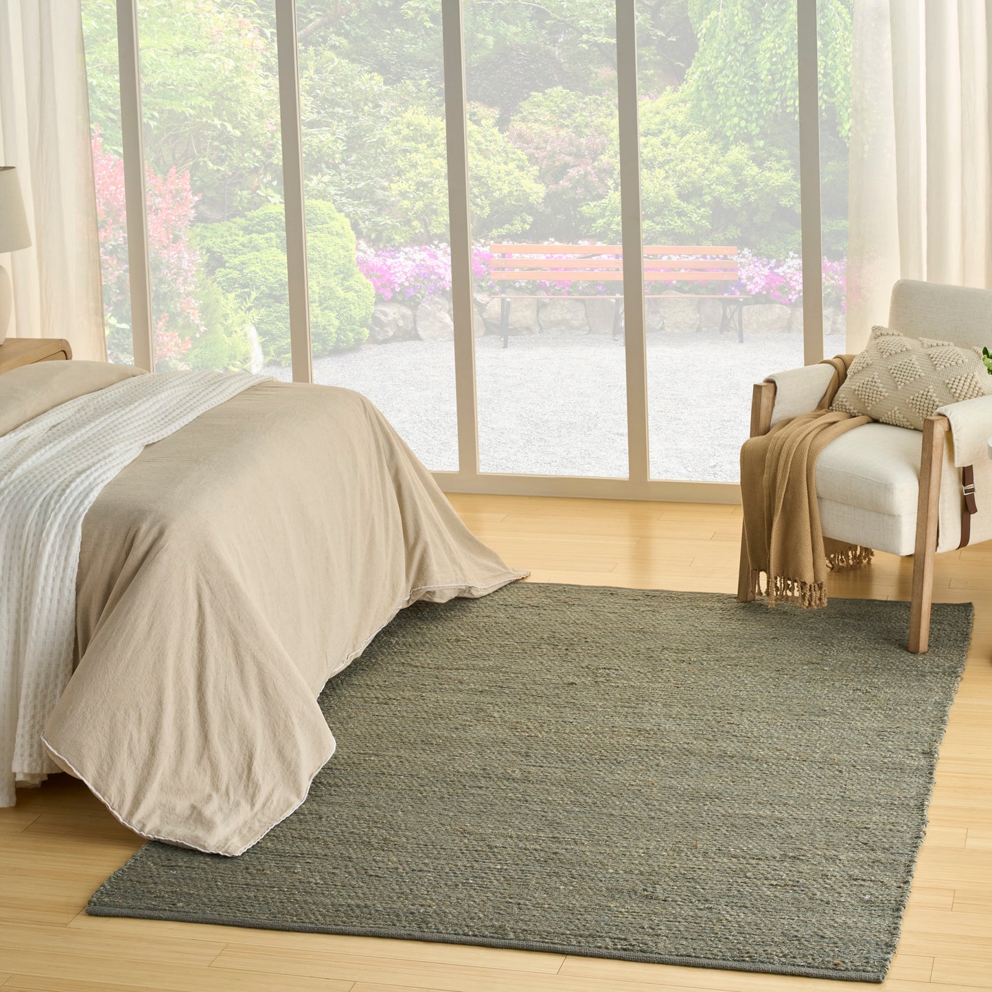 Bedroom with beige bedding, gray rug, wooden floor, armchair with a throw by glass patio doors opening to a garden.