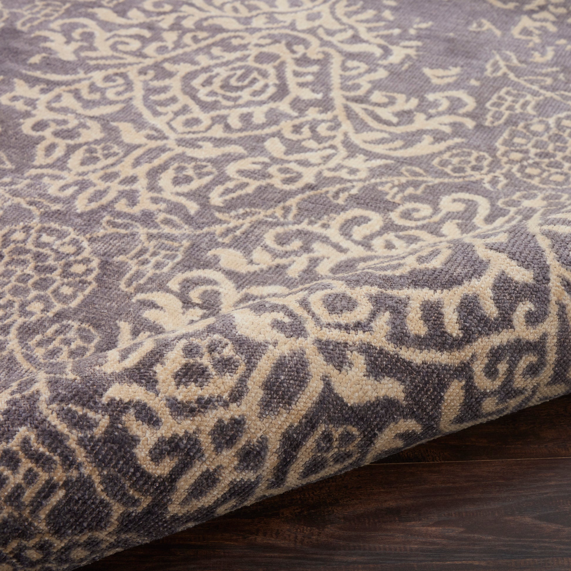 Gray damask-patterned area rug with ivory motifs, folded edge on dark wood floor.